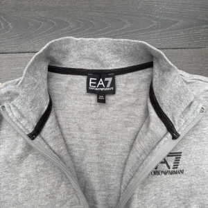 Grå mjukisbyxor från EA7 Emporio Armani + tröja - Grå mjukisbyxor från EA7 Emporio Armani med svart logga på vänster lår. Byxorna har resår i midjan, raka ben och sidofickor. Tillverkade i mjukt bomullsmaterial som är perfekt för chill eller träning.