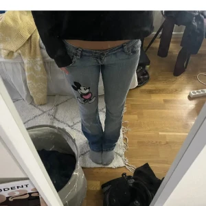 Blå jeans med Mickey Mouse tryck - Säljer ett par ljusblå jeans med coolt Mickey Mouse tryck på både framsidan och bakfickan. Byxorna har låg midja och klassisk femficksdesign. Passformen är straight med lätt utsvängda ben. Perfekt för dig som vill sticka ut med en unik detalj.