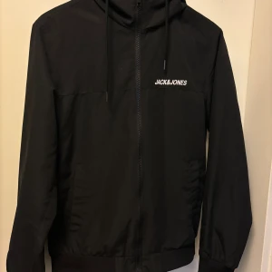 Svart vindjacka Jack & Jones XS - Svart vindjacka från Jack & Jones i storlek XS. Jackan har huva med snörning, dragkedja framtill och två sidofickor. Stilren design med logotyp på bröstet och ribbade muddar vid ärmslut och nederkant. Perfekt för dig som gillar enkel och clean look.