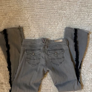 Grå Lågmidjade spets jeans - Snygga grå jeans från Super Soin i bootcut passform och superlow midja. Byxorna har svarta volangdetaljer längs utsidan av benen och dragkedjor vid bensluten. Perfekta för dig som vill sticka ut med en unik look., sitter som en m