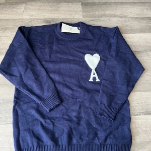 Mörkblå stickad tröja från Ami - Snygg mörkblå stickad tröja från Ami med vit hjärtdetalj och bokstaven A på bröstet. Tröjan har rund hals, ribbade muddar och lång ärm. Perfekt för dig som gillar stilrena plagg med en twist.