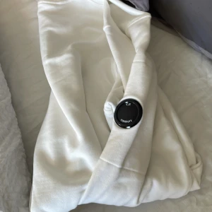 Vit sweatshirt från CP Company - Vit sweatshirt från CP Company med klassisk rund hals och ribbade muddar vid ärmslut och nederkant. Tröjan har en ikonisk svart linsdetalj på ena ärmen och är tillverkad i mjuk bomull. Perfekt för dig som gillar clean och stilren streetwear.