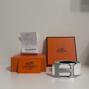 Vit H skärp med H-spänne - Exklusivt vitt H bälte i skinn med ett stort, stilrent H-spänne i silverfärgad metall. Bältet har en klassisk, rak form och synliga vita sömmar längs kanterna. Perfekt för dig som vill ha en lyxig accessoar med tydlig designerstil.