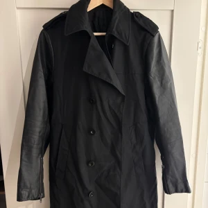 Svart trenchcoat från tiger of sweden med skinnärmar - Svart trenchcoat från tiger of sweden med coola skinnärmar och klassisk krage. Jackan har dubbelknäppning, axelklaffar och dragkedjor vid ärmsluten. Tyg tillverkat i Italien, insida med kontrastficka. Perfekt för dig som gillar stilrent! Pris går att diskutera! 