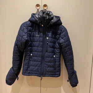 Uhip jacka - Marinblå pufferjacka från Uhip med huva, dragkedja framtill och två sidofickor med dragkedja. Den här även uppvikbara ärmar!