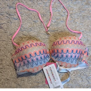 Färgglad bikini-bh med vågigt mönster - Säljer en bikini-bh med vågigt mönster i rosa, blått och beige. Kuporna är vadderade och har en balconette-form. Axelbanden är ljusrosa och justerbara, och bh:n stängs med ett guldfärgat spänne bak. Perfekt för strandhäng i sommar!