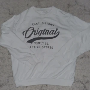 Vit sweatshirt från H&M med tryck - Vit sweatshirt från H&M med svart texttryck 'Original East District Supply Co. Active Sports' på bröstet. Tröjan har rund hals, långa ärmar och ribbade muddar vid ärmslut och nederkant. Perfekt för en sportig och avslappnad stil.