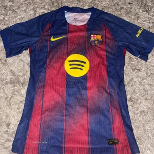 FC Barcelona Gavi #6 matchtröja Nike - Säljer en officiell FC Barcelona fotbollströja från Nike med Gavi #6 på ryggen. Tröjan har klassiska blå och röda ränder, gula detaljer och klubbmärke på bröstet. Tillverkad i lätt funktionsmaterial med korta ärmar och sponsorer som Spotify och UNHCR tryckta på.