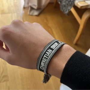 Christian Dior svart/vitt armband - Säljer ett snyggt armband från Christian Dior i svart och vitt med tydlig logga framtill. Armbandet är vävt i tyg och har tofsar och knytning baktill för en cool och avslappnad look. Perfekt accessoar för att lyfta din outfit.