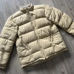 Beige pufferjacka från Nike - Säljer en beige pufferjacka från Nike med svart broderad swoosh på bröstet och ärmen.   Jackan har hög krage, dragkedja framtill och två sidofickor. Materialet är mjukt och perfekt för kalla dagar. Enkel och clean design som funkar till allt. Kan tänka mig att gå ner i pris vid snabb och smidig affär!  ✅ Skick: 10/10 ✅ Storlek: XL ✅ Mått: 80x67cm