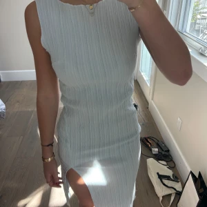 Ljus ribbad ärmlös klänning med slits - Säljer en ljus, ärmlös klänning med ribbad struktur och hög slits på sidan. Klänningen har en kroppsnära passform och är tillverkad i ett lätt, mjukt material som faller snyggt. Oanvänd.