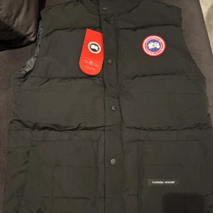 Svart dunväst från Canada Goose - Svart dunväst från Canada Goose med klassisk logotyp på bröstet och märkning på fickan. Västen har tryckknappar framtill, två stora fickor och quiltad design. Perfekt för kalla dagar och har en stilren look som funkar till många outfits.