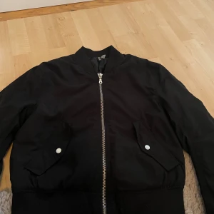 Svart bomberjacka med dragkedja - Säljer en svart bomberjacka med silvrig dragkedja framtill och två fickor med tryckknappar. Jackan har ribbade muddar vid midja och ärmslut samt en klassisk bomberkrage. Materialet känns lätt och smidigt, perfekt för lager-på-lager-stil. Köpte för 300 men säljer för 200 kr 