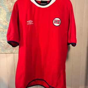 Norge Umbro 2000/02 shirt, storlek XL - Riktigt snygg röd fotbollströja från Umbro med Norges landslagsemblem på bröstet. Tröjan har vita och mörkblå detaljer vid krage och ärmslut. Tillverkad i lätt och ventilerande polyester, perfekt för match eller träning.