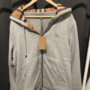  Grå Burberry Check Zip hoodie - Grå hoodiejacka från Burberry med klassiskt rutigt foder i beige, svart och rött. Jackan har dragkedja, snörning i huvan och broderad Burberry-logga på bröstet. Tillverkad i mjuk bomull- och polyesterblandning, perfekt för en chill men stilren look. Storlek L, passar även Medium för en något oversized fit.  🖤 Notera: Produkten postas endast – ingen mötesaffär. 📌 Läs gärna bio på min Plick-profil innan köp. 