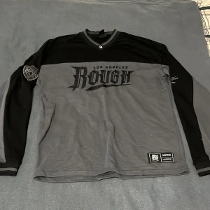 Grå och svart oversized tröja Black Squad - Säljer en oversized långärmad tröja från Black Squad i grått och svart med coolt 'Los Angeles Rough' tryck på bröstet och patch på ärmen. V-ringad krage med randiga detaljer och flera broderade emblem. Perfekt för dig som gillar streetwear.