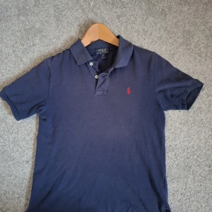 Marinblå pikétröja från Ralph Lauren - Klassisk marinblå pikétröja från Ralph Lauren. Nypris 800 kr.                                                        Röd broderad logga på bröstet. Tröjan har korta ärmar, krage och knäppning med två knappar. Perfekt för en stilren och avslappnad look.
