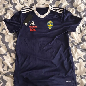 Svenska landslaget fotbollströja Adidas - Säljer en officiell blå fotbollströja från Svenska landslaget, Adidas. Tröjan har korta ärmar, vit V-ringad krage och vita detaljer på axlarna. Materialet är lätt och andas, Aeroready-teknologi. Tryck med SvFF-emblem, Bauhaus och ICA på bröstet.