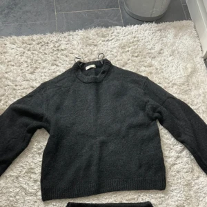 Svart stickad tröja med rund hals - Mysig svart stickad tröja med rund halsringning och ribbade muddar vid ärmslut och nederkant. Tröjan har en klassisk, enkel design och är långärmad, perfekt för kyligare dagar. Passformen är normal och materialet känns mjukt och värmande.