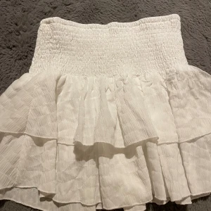 Vit volangkjol med smock - Supersöt vit kortkjol med bred smock i midjan och flera lager volanger. Kjolen har en lätt strukturerad yta och är perfekt för sommarens alla tillfällen. Den är luftig och har en riktigt fräsch vibe. Storlek M/S passar nog L också.