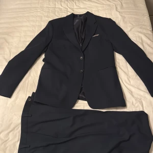 Mörkblå kostym från Zara - Säljer en stilren mörkblå kostym från Zara med klassisk tvåknäppt kavaj och matchande byxor. Kostymen har slät finish, smala slag och innerfoder i satin. Perfekt för dig som vill ha en clean och modern look.