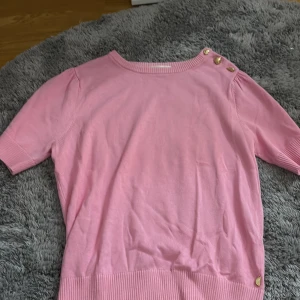 Rosa topp med hjärtknappar Fabienne Chapot - Säljer en ljusrosa topp från Fabienne Chapot i storlek XS. Toppen har korta ärmar, ribbade muddar och tre guldiga hjärtformade knappar vid axeln som ger en gullig detalj. Perfekt för vår och sommar, och materialet känns mjukt och skönt mot huden.