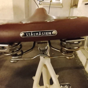 Stålhästen Damcykel - Elegant vit Stålhästen damcykel med klassisk design och brunt sadel och handtag. Cykeln ser välvårdad ut med retrodetaljer och bekväm fjädrad sadel. Inga synliga skador eller slitage, perfekt för stadscykling och vardagsbruk.