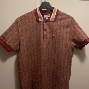 Rutig röd kortärmad skjorta från Lacoste - Snygg röd skjorta från Lacoste med rutigt mönster och klassisk krokodillogga på bröstet. Skjortan har korta ärmar, krage med kontrastfärg och knappar framtill. Materialet känns mjukt och andas, perfekt för dig som gillar stilrena detaljer.