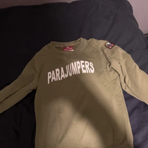 Olivgrön tröja från Parajumpers - Snygg olivgrön långärmad tröja från Parajumpers med stort vitt tryck på bröstet och patch på ena ärmen. Rund hals och mjukt bomullsmaterial gör den skön att bära. Perfekt för dig som gillar streetwear och vill ha en clean look. nypris 1800