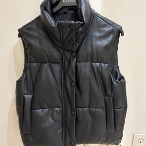  Ny Svart quiltad dunväst från Zara - Svart quiltad dunväst från Zara med hög krage och tryckknappar framtill. Västen har en stilren look med släta paneler och två fickor på sidorna. Materialet är syntet med skinnliknande finish som ger en cool vibe. Perfekt att slänga över en hoodie för extra edge.