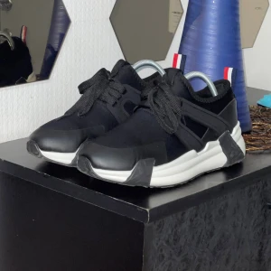 Svarta sneakers från Moncler - Snygga svarta sneakers från Moncler med vit platt sula och svarta snören. Skorna har en modern design med detaljer i syntet och textil, samt en röd, vit och blå flik bak. Yttersulan har ett unikt mönster för extra grepp. Perfekt för dig som gillar stilrena och sportiga skor.