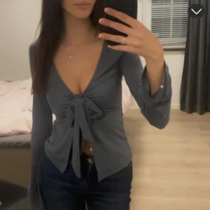 Grå omlottopp med knyt från Gina Tricot - Från Gina Tricot i storlek XS. Lång ärm, djup v-ringning och snygg knytning framtill. Använd 1 gång endast