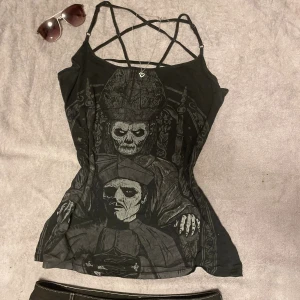 Svart Ghost linne med tryck och strappy rygg - Svart linne med smala axelband och coolt strappy mönster i ryggen. Stort tryck framtill med motiv av Papa Emeritus från Ghost och bandets logga broderad på ryggen. Materialet är mjuk bomull, passformen är tight och toppen är ärmlös.