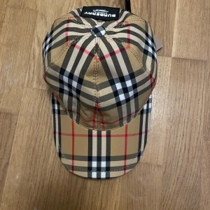Burberry rutig keps beige/svart M - Snygg Burberry keps i klassiskt rutigt mönster med beige, svart, vit och röda toner. Kepsen har böjd skärm, justerbar rem och tydlig Burberry-logga baktill. Materialet är mjukt och slitstarkt, perfekt för dig som vill ha en ikonisk streetstil.