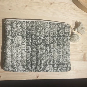 Quiltad datorväska med blommigt mönster - Trendig quiltad datorväska i grön/grått och vit mönster. Fodralet har dragkedja upptill och två stora tofsar som detalj, helt ny.😊💕 Köpt för 249