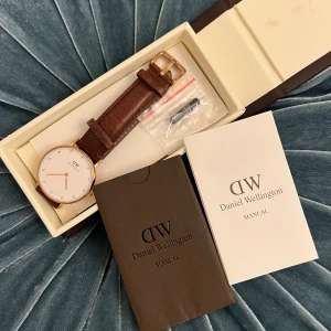 Daniel Wellington klocka med brun läderrem - Snygg klocka från Daniel Wellington med rund vit urtavla, guldiga detaljer och kristallmarkeringar. Remmen är i brunt skinn med guldspänne. Stilren och klassisk design som passar perfekt till dig som gillar enkla men eleganta accessoarer. Klockan är i gott skick kosmetiskt. Batteriet fungerar inte just nu, så den behöver ett nytt för att starta.