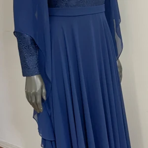 Marinblå långklänning med spetsdetaljer - Elegant marinblå långklänning med vackert spetsmönstrad överdel och långa, flödande ärmar. Kjolen är vid och faller snyggt ner till golvet. Perfekt för dig som vill ha en klassisk och stilren look med extra detaljer på ärmar och överdel.