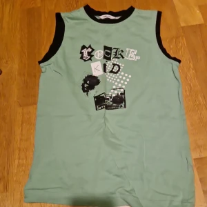 Mintgrön ärmlös topp Rocker Kid Lindex - Cool mintgrön ärmlös topp från Kids by Lindex med svart tryck och texten 'Rocker Kid' framtill. Svarta kantband vid hals och ärmhål ger en snygg kontrast. Perfekt för dig som gillar en rockig stil och vill sticka ut lite extra.