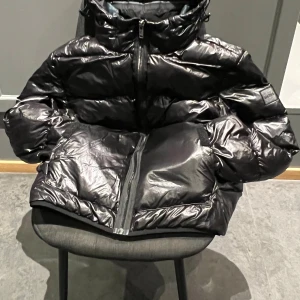 Svart pufferjacka från BOSS - Säljer en svart, glansig pufferjacka från BOSS med hög krage och tydlig logotyp på ärmen. Jackan har dragkedja framtill, två sidofickor och en loose passform. Perfekt för kalla dagar med sin tjocka vaddering och stilrena look.