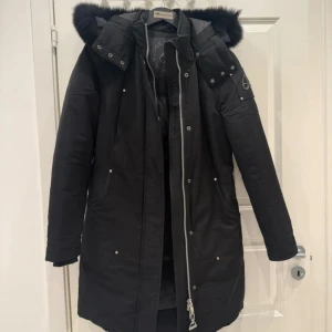 Svart parkas från Moose Knuckles - Svart parkas från Moose Knuckles med fluffig pälskantad huva och silverfärgade detaljer. Jackan har lång passform, dragkedja framtill, tryckknappar och logotyp på ärmen. Perfekt för kalla dagar med sin varma och stilrena look.