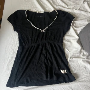 Svart Odd Molly blus med spetsdetaljer - Supersöt svart blus från Odd Molly med ribbad struktur och v-ringning. Dekorerad med vit spets runt hals och ärmslut samt en liten rosett framtill. Kortärmad och med Odd Molly-logga i form av en fjäril på ena ärmen. Perfekt för dig som gillar detaljer!
