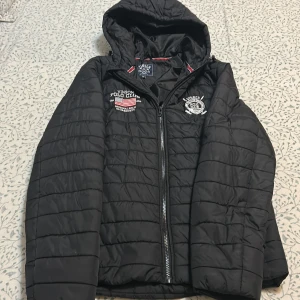 Svart pufferjacka från Vinson Polo Club - Svart pufferjacka från Vinson Polo Club med broderade detaljer på bröstet, inklusive klubbens logga och amerikansk flagga. Jackan har huva, dragkedja framtill och quiltad design. Perfekt för dig som gillar sportig och clean stil.