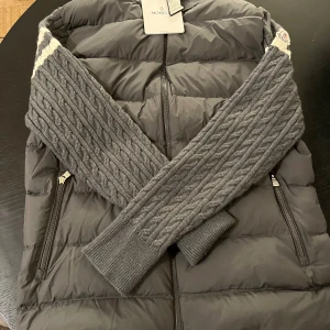Grå Cardigan från Moncler - Snygg grå Cardigan från Moncler med stickade kabelmönstrade detaljer och dragkedja framtill. Jackan har två sidofickor med dragkedja och Moncler-logga på axeln. Materialmix av dun och stickad ull ger en unik look. Perfekt för lager-på-lager under kyliga dagar. Storlek är 3xXL men sitter som en vanlig XL. 
