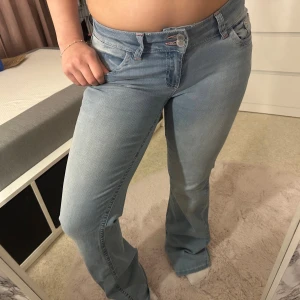 Ljusblå bootcut jeans från Nelly - Säljer ett par ljusblå jeans från Nelly med bootcut passform. Jeansen har klassisk femficksdesign, dubbla knappar i midjan och snygga rosa broderade detaljer på bakfickorna. Tillverkade i mjuk bomullsdenim med lite stretch för extra komfort.