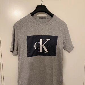 Grå Calvin Klein Jeans t-shirt - Snygg grå t-shirt från Calvin Klein Jeans med stort svart tryck och vit CK-logga på bröstet. Klassisk rund hals och korta ärmar. Tillverkad i mjuk bomull för en skön känsla. Perfekt för en clean och stilren look.
