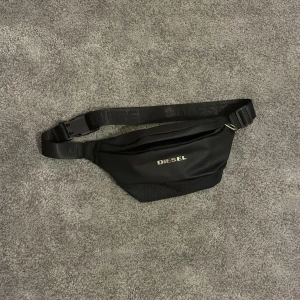 Svart magväska från Diesel - Snygg svart magväska från Diesel med justerbart band och diskret logga i silver framtill. Väskan har dragkedja och är tillverkad i slitstark polyester. Perfekt för att bära det viktigaste och hålla stilen enkel och clean.