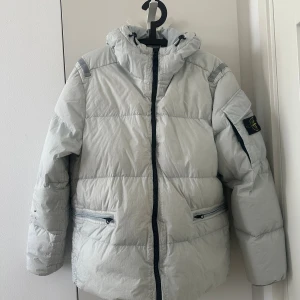 Ljusgrå dunjacka från Stone Island - Säljer en fet ljusgrå dunjacka från Stone Island med huva och svarta detaljer. Jackan har dragkedja framtill, två fickor med dragkedja och klassisk patch på ärmen. Perfekt för kalla dagar och riktigt snygg streetstil.