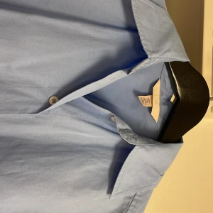 Ljusblå oversized skjortklänning från H&M - Oversized skjortklänning i ljusblå bomull från H&M med klassisk krage och vita knappar hela vägen ner. Skjortan har lång ärm med knapp vid manschetten och en loose passform som ger en chill vibe. Perfekt för dig som gillar enkel och stilren design. Observera, en liten fläck vid både ärmsluten.