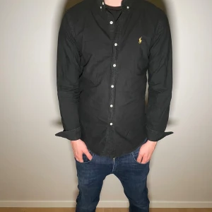 Ralph Lauren skjorta - Svart skjorta från Polo Ralph Lauren med vita knappar. Broderad gul logga på bröstet. Skjortan är långärmad och har en stilren look.