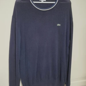Marinblå stickad tröja Lacoste XXL - Snygg marinblå stickad tröja från Lacoste i storlek XXL. Tröjan har rund hals med vit och blå randig kant, klassisk krokodillogga på bröstet och långa ärmar. Perfekt för dig som gillar stilrena och sportiga plagg med premiumkänsla.
